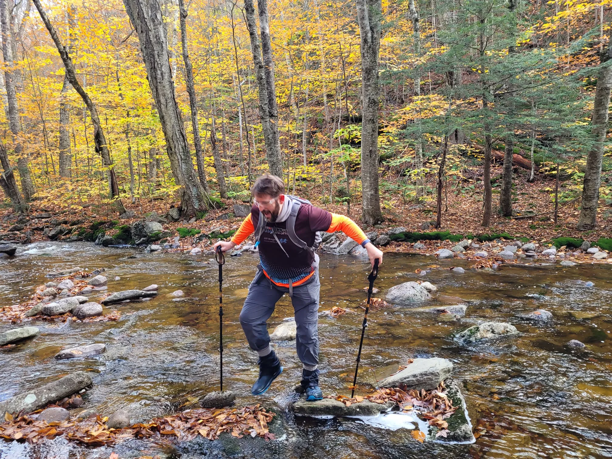 A man (Adam) with a short beard, dressed for hiking, using hiking poles to cross a shallow but wide rocky stream in a forest. This picture is zoomed closer to the figure and he is caught in motion, balancing mid-step as he carefully goes for the next stream rock foothold.